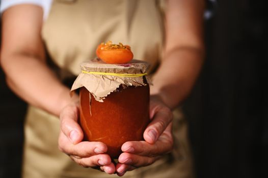 Details: Hands of a woman confectioner in a beige chef apron, handing a jar of homemade jam with juicy ripe red summer sunny apricot broken in halves on the craft lid. Canning, preserved sweet food.