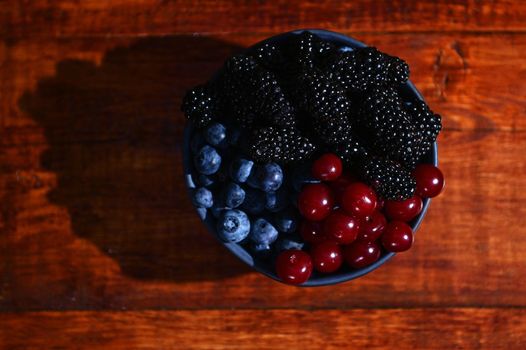 Flat lay. Blue ceramic bowl of fresh, ripe and ready-to-eat organic wild berries- blueberries, blackberries and cherries from and organic farm, on a wooden background. Copy ad space for text. Banner