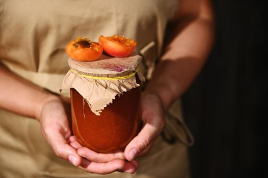 Details: Hands of a woman confectioner in a beige chef apron, holding a jar of homemade jam with juicy ripe red summer sunny apricot broken in halves on the craft lid. Canning, preserved sweet food.