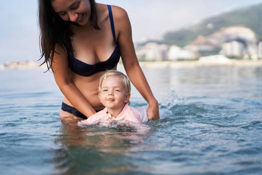 Little girl swims with her tongue out, supported by her mother. High quality photo
