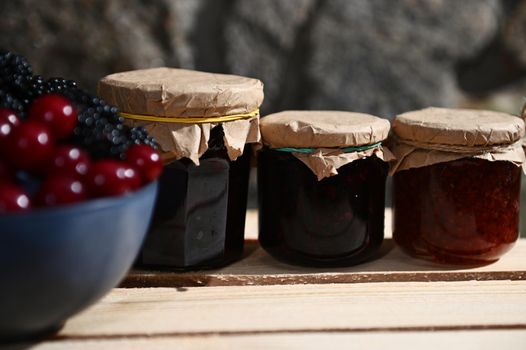 Assortment of homemade berry jam from red currant, cherry and blackcurrant on a wooden surface, with a blurred blue bowl with fresh ripe berries in the foreground. Canned food, canning for winter