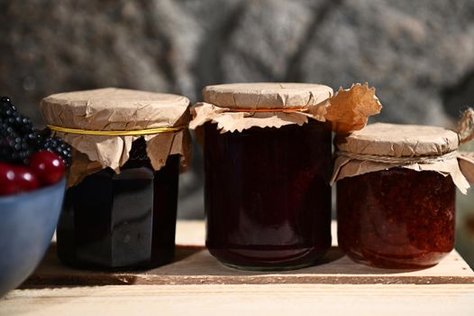 Collection of homemade berry jam from red currant, cherries and blackcurrant on a wooden surface, with a blurred blue bowl with fresh ripe berries in the foreground. Canned food, canning for winter