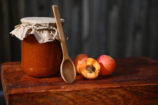 Still life. A glass jar of homemade jam, with craft paper on the lid tied with rope, halves of ripe ready-to-eat red sunny apricots and wooden spoon on rustic wood surface. Canning, sweet preserves.