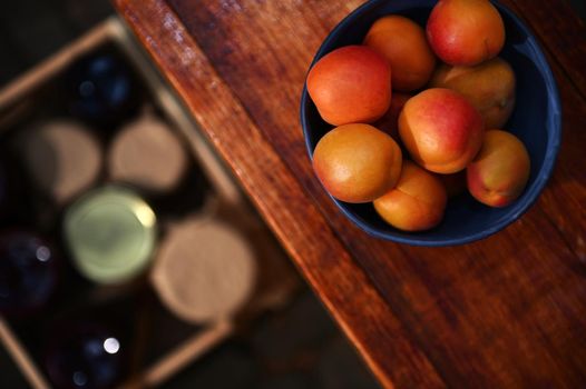 Soft focus on ripe, ready-to-eat apricots in a blue ceramic bowl on a wooden surface against a blurry wooden box with jam jars on the floor. Organic farm and canning concept