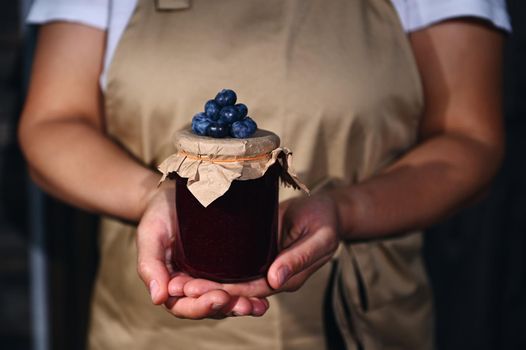 Canning, preserved food concept for winter season. Focus on a jar of blueberry jam with fresh berries on the craft lid, in the hands of a woman confectioner wearing a chef apron. Close-up.