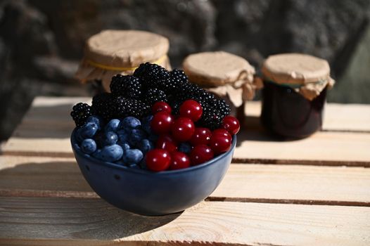 Close-up of a blue ceramic bowl with ripe cherry berries, blueberries and blackberries in a wooden box against the background of blurry assorted fruit jams in jars. Organic farm and canning concept