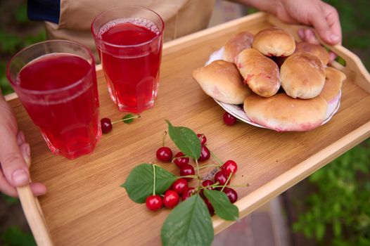 Overhead view of the hands of a housewife holding a serving tray with freshly baked cherry pies and drinking glasses with refreshing compote from cherries, standing in the backyard of a private house