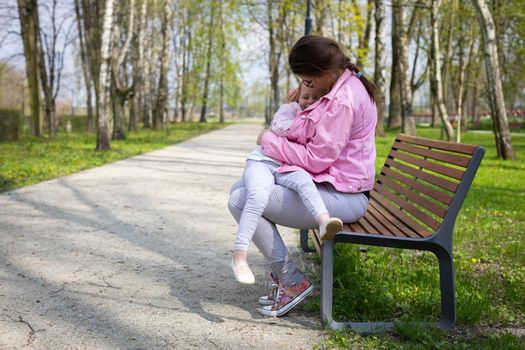A young mother with her four-year-old daughter are sitting on a bench in a city park. The little girl is cuddling with her mother. A spring sunny day.