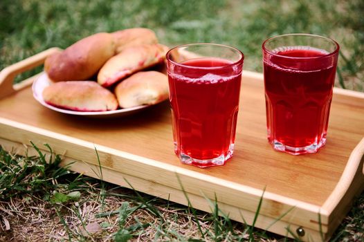 Delicious homemade cherry pies and two drinking glass of berry compote on a tray on the grass in the garden. Food and drink concept