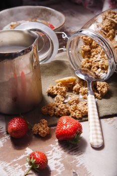 Homemade scattered granola in glass jar with fresh strawberry and milk on wooden table, selective focus