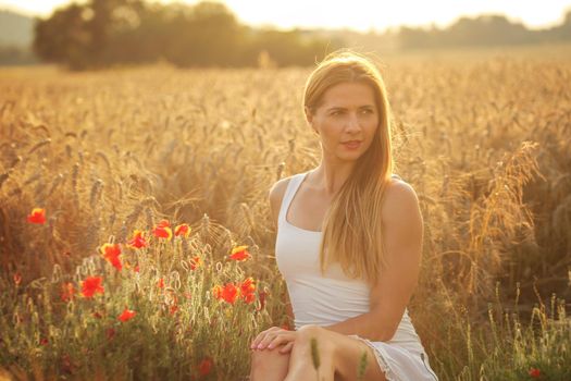 Young woman in white dress, sitting in wheat field, red poppy near, lit by sunset light.