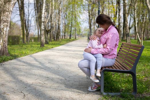 On a spring morning in the park, a mother and her daughter sit on a bench. The four-year-old girl is smiling. The mother holds the child in her lap on a sunny day.