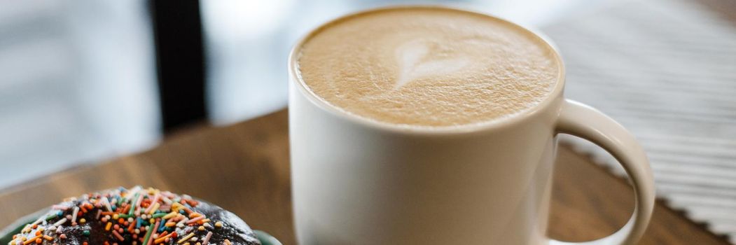 Coffee with a drawn heart and milk on a wooden table in a coffee shop. Pink donut with scattering on the table next to the coffee. Web banner.