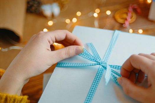 Women's hands tie blue ribbon bow on white gift box. Surprise for Birthday or Christmas. New Year's mood. Garland lights and decorations in background.