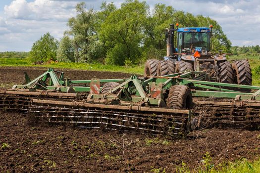 Blue New Holland tractor with double wheels pulling disc harrow with roller basket at hot sunny day in Tula, Russia - June 4, 2022