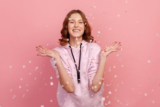 Cheerful young woman rejoicing falling heart shaped confetti, catching glitter with open palms and smiling at camera, girl celebrating valentines day. Indoor studio shot, isolated on pink background