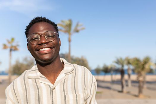 Portrait of young happy and smiling black man wearing glasses outdoors. Background with palm trees. Copy space. Lifestyle and vacation concept.
