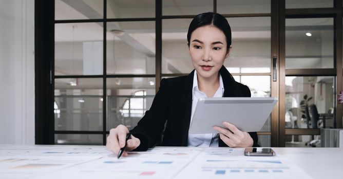 Attractive and charming young Asian businesswoman or financial worker in formal suit using tablet and working with her financial report in the office..