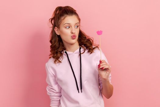 Portrait of funny teen girl with ponytail and pout lips wearing hoodie looking at paper party props, having fun on holiday, carnival accessories. Indoor studio shot, isolated on pink background