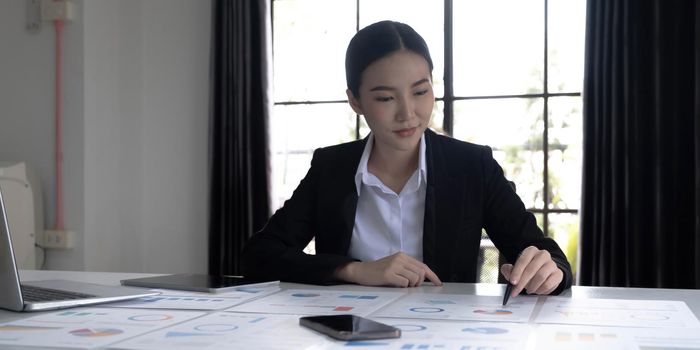 Young Asian businesswoman analyzing report pointing at graph holding a pen using a tablet laptop placed at the office..