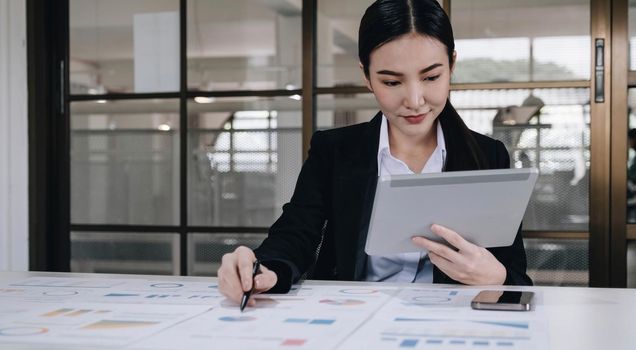 Attractive and charming young Asian businesswoman or financial worker in formal suit using tablet and working with her financial report in the office..