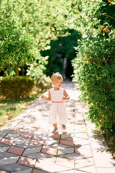 Little girl walks along the paving stones near a green bush in the park. High quality photo