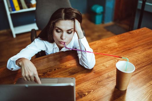 Business woman working laptop computer drinking coffee through straw. Busy young woman works at office workplace. Crisis concept. Tinted image.