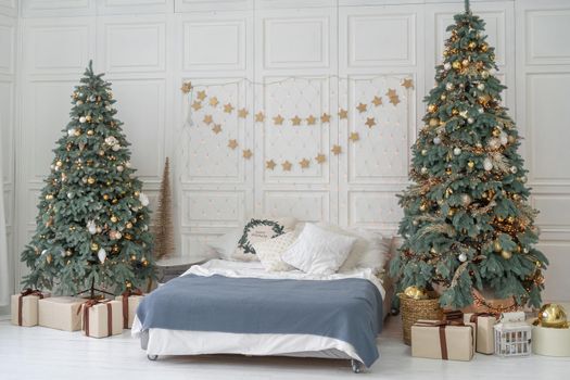 New Year bedroom interior. Bed with blue bedspread stands between two Christmas trees decorated with balls and tinsel. Above bed hangs garland with stars. New Year's gifts stand under Christmas trees.