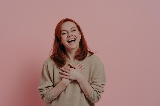 Ha-ha. Delighted red-haired funny woman laughing out loud, dressed in beige sweater, holding both hands on chest and smiling cheerfully, posing against pink background with copy space for text