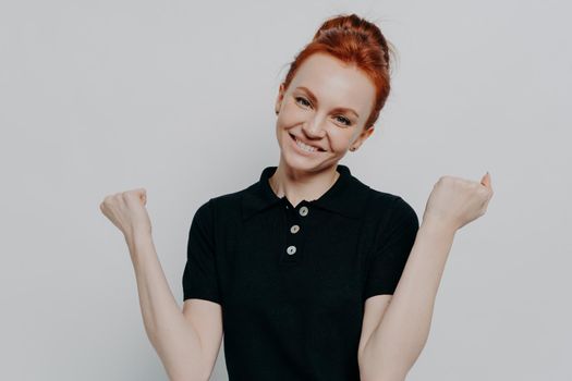 Victory concept. Studio shot of happy caucasian redhead woman clenching fists and smiling happily, excited ginger female in black casual t shirt achieving goal, isolated over grey background