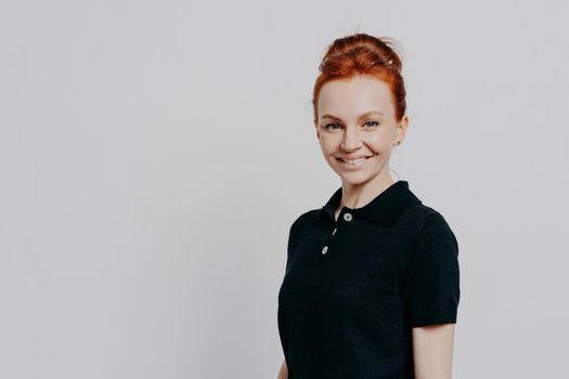 Headshot of young positive millennial red haired woman looking at camera with charming smile, posing isolated over grey background with copy space, being satisfied and overjoyed. Positive emotions