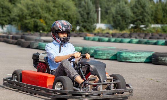 A girl or a woman in a hard hat rides a go-kart on a special track fenced with rubber wheels. Active recreation and sports on transport. Preparation and training for competitions. 