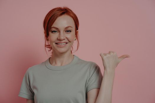 Horizontal shot of cheerful redhead woman with happy facial expression indicating with finger at blank copy space, female pointing with thumb while advertising product. Advertisement concept