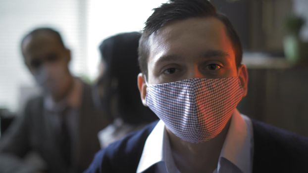 White Collar Worker In Protective Mask Looking At Camera Seriously, Young Man Backlit By Red Neon Light On The Right, His Colleagues Are Talking On Blurred Background