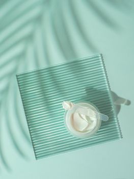 Cosmetic cream moustirizer, transparent ribbed acrylic plate, tropical palm leaf shadows on blue background. Open round glass jar,aesthetic swirls cream,spatula. Top view flatlay. Copy space. Vertical
