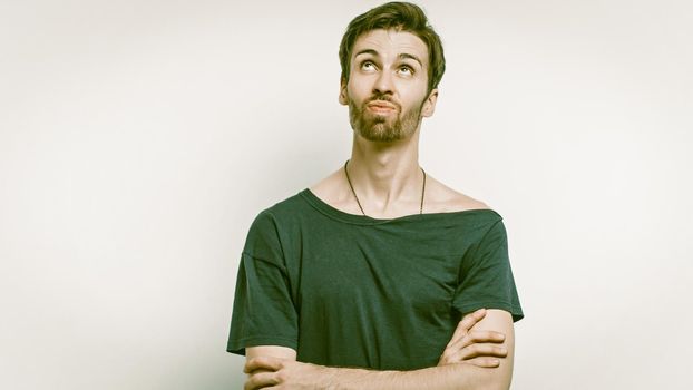 Bearded Man Grimaces Looking Up, Frontal View Of Caucasian Male Waiting For The Manna Of Heaven, Young Handsome In Black Clothes Stands With Crossed Arms On Gray Background, Copy Space On The Left