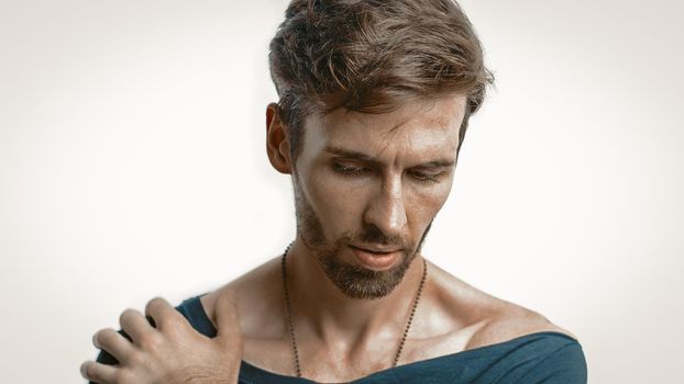 Close-Up Portrait Of A Handsome Pensive Man, A Caucasian Man In A Black T-Shirt With A Wide Neckline Lowered His Gaze Down With His Hand On His Shoulder, Emphasized Focus On Male Face