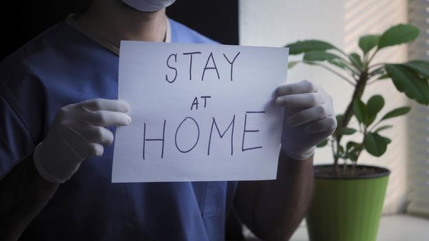 Doctor Or Nurse Shows Sign To Stay At Home, Male Medic In Blue Uniform Demonstrates Inscription On Piece Of Paper, Medical Worker Stands Near Window In His Office, Quarantine Concept