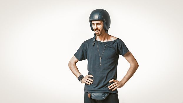 Cute Motorbiker In Protective Helmet Standing Akimbo, Male Portrait Of Caucasian Man In Black Sports Clothing Looks At Left Side Toothy Smiling, Isolated On White, Copy Space At Left Side