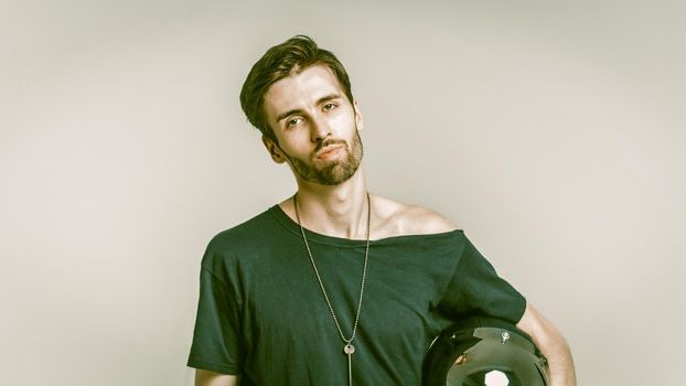 Confident Motorcyclist Looking At The Camera With His Head Bowed To The Side, Front View Of Stylish Bearded Man In Black Wide-Neck T-Shirt Holding A Safety Helmet With Hand, Cut Out On Gray Background