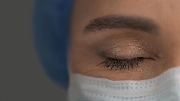 Tired Female Medic In Protective Uniform Closed Her Eyes, Young Woman Exhausted By Fighting Coronavirus Epidemic, Close Up Of Female Doctor's Closed Eye, Copy Space At Left Side On Grey Background