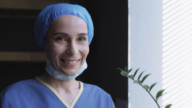 Toothy Smiling Female Medic Looks At Camera, Happy Doctor Or Nurse In Blue Uniforme Rejoices Standing Near Window In Her Office, Quarantine Concept