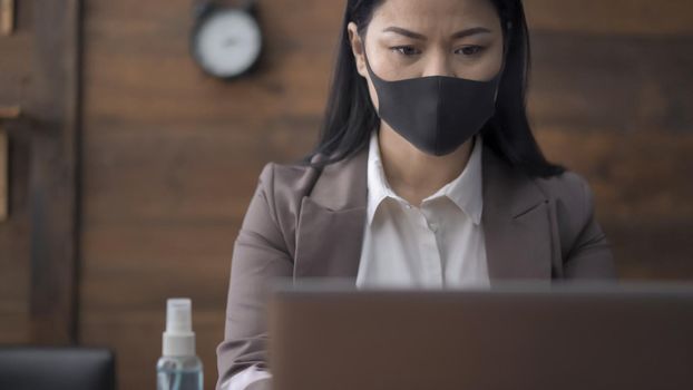 Young Woman In Protective Mask Works With Computer Sitting At Table With Sanitizer Spray On It, Close Up Portrait Of Business Woman Working In Office Despite Quarantine, Virus Outbreak Concept