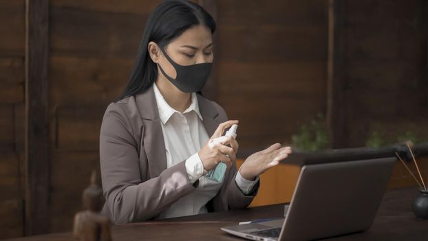 Business Woman In Mask Disinfects Hands Sitting At Office Desk, Asian Brunette Woman Sprinkles With Sanitizer Spray On Hands For Disinfection While Working With Computer In Office During Quarantine