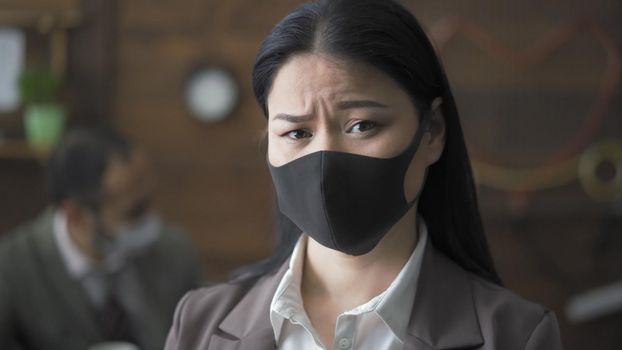 Worried Asian Woman Looking At Camera Against Background Of Wooden Wall Of Cozy Office, Portrait Of Female Office Workers In Black Medical Mask Working Despite Quarantine, Coronavirus Pandemic Concept