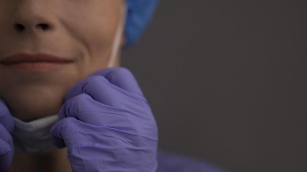 Female Doctor Or Nurse Adjusts Protective Mask, Selective Focus On Woman's Hand In Blue Rubber Gloves, Copy Space At Right Side On Grey Background, Pandemic Concept