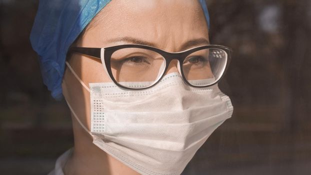 Tired Female Doctor In Mask And Glasses Looking At Window, Young Woman Is Forced To Work Hard During An Epidemic, Close Up Of Female Face In Protective Cap And Mask, Quarantine Concept