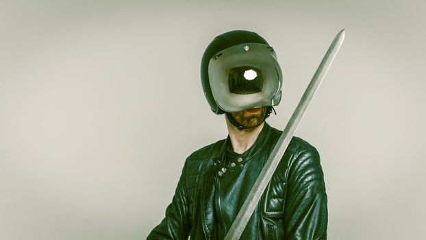 Mystical Knight In A Black Holding Sword, Caucasian Man In Black Leather Jacket And In Helmet With Glass Down, Holds A Double-Edged Sword In Front Of Himself, Studio Shot On A Gray Background