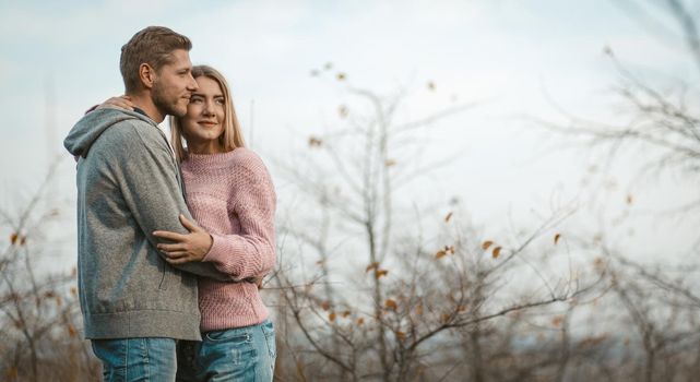 Loving Couple Is Standing In An Embrace In Nature, Young Woman And Man Are Hugging While Admiring The Sunset Against Sky And Autumn Trees, Copy Space At Right Side