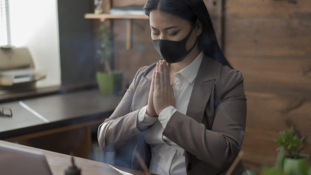 Asian Woman In Mask Praying In Isolation During Epidemic, Business Woman Prays In Office With Closed Eyes And Putting Her Palms Together While Sitting At Desk, Coronavirus Outbreak Concept